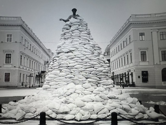 A black and white photograph by Eddy van Wessel of the Duke of Richelieu monument in Odessa, Ukraine, during the war. The statue is almost entirely encased in a massive, towering pyramid of white sandbags for protection. Only the bronze head and one gesturing arm of the figure emerge from the top of the stack. The monument is situated in a paved square, flanked by large neoclassical buildings under a grey sky.