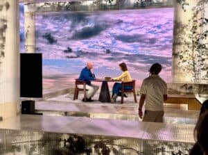 A photograph from the VPRO television programme Zomergasten, showing Dutch writer and Protect Ukraine ambassador Tommy Wieringa being interviewed by presenter Janine Abbring. Wieringa sits on the left wearing a blue shirt and white trousers, facing Abbring on the right, who wears a yellow top and teal trousers. They are seated at a small round table in a modern studio set featuring translucent ribbed panels and a large backdrop displaying a cloudy sunset sky. A crew member is partially visible in the right foreground.
