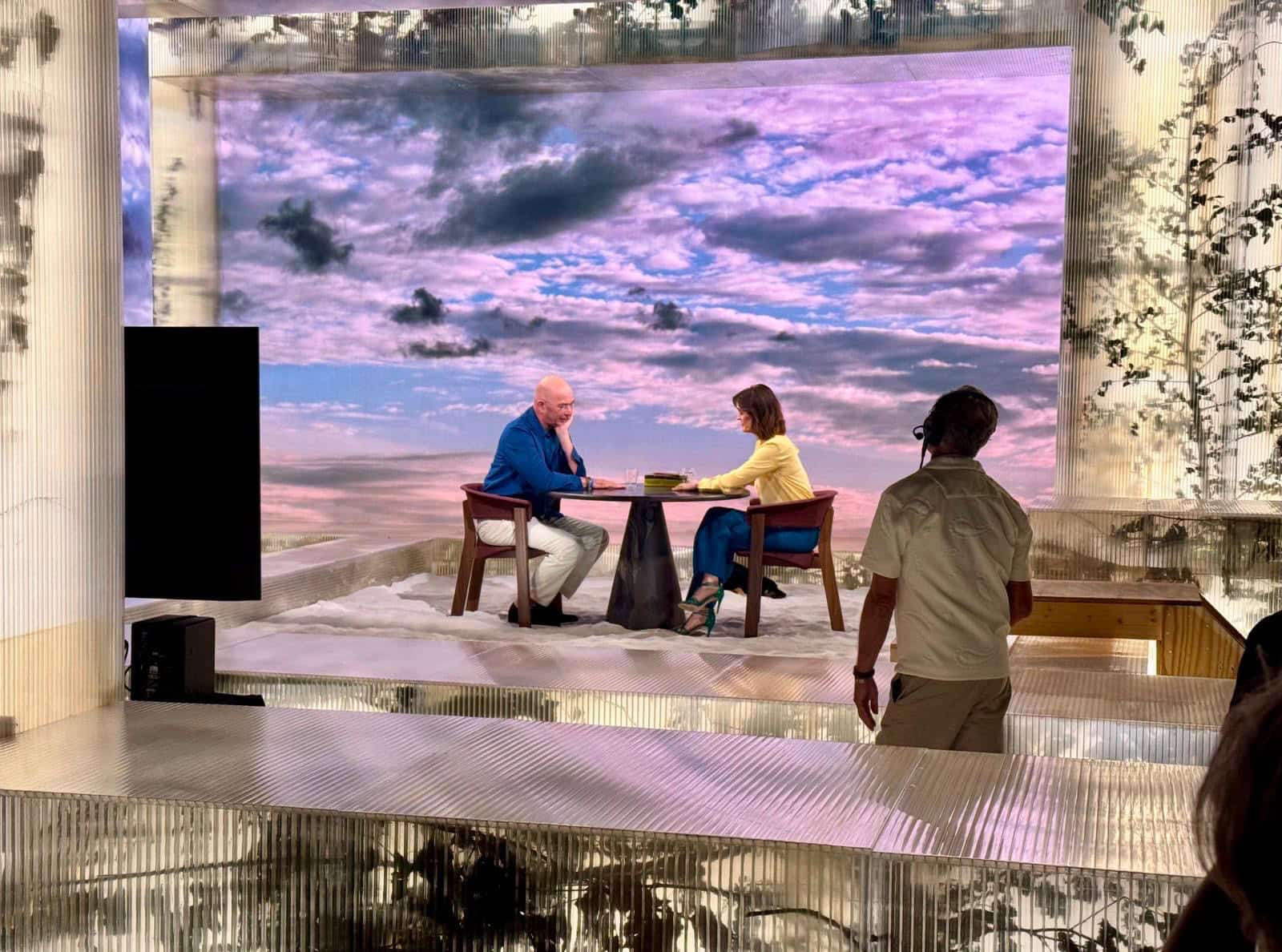 A photograph from the VPRO television programme Zomergasten, showing Dutch writer and Protect Ukraine ambassador Tommy Wieringa being interviewed by presenter Janine Abbring. Wieringa sits on the left wearing a blue shirt and white trousers, facing Abbring on the right, who wears a yellow top and teal trousers. They are seated at a small round table in a modern studio set featuring translucent ribbed panels and a large backdrop displaying a cloudy sunset sky. A crew member is partially visible in the right foreground.